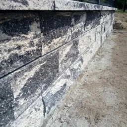 Niski mur oporowy z szarej kostki łupanej, widoczny z bliska, z siatką ogrodzeniową w tle, na nierównym terenie.