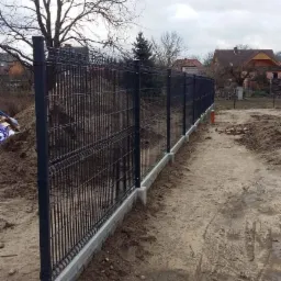 Długa, metalowa, panelowa ogrodzenie w kolorze grafitowym, osadzone na betonowej podmurówce z widocznymi słupkami, teren wokół ogrodzenia częściowo wyrównany, w tle domy jednorodzinne i drzewa bez...