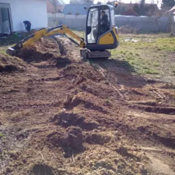 Żółto-szara mini koparka Wacker Neuson podczas wykopu wąskiego rowu w ziemi, widoczny operator w kabinie, w tle budynek w trakcie budowy i osoba pracująca łopatą.