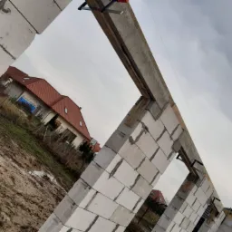 Wznoszone ściany domu z betonu komórkowego Ytong, widoczne zbrojenie nadproża i zaciski stolarskie, w tle dom z czerwoną dachówką i pochmurne niebo.