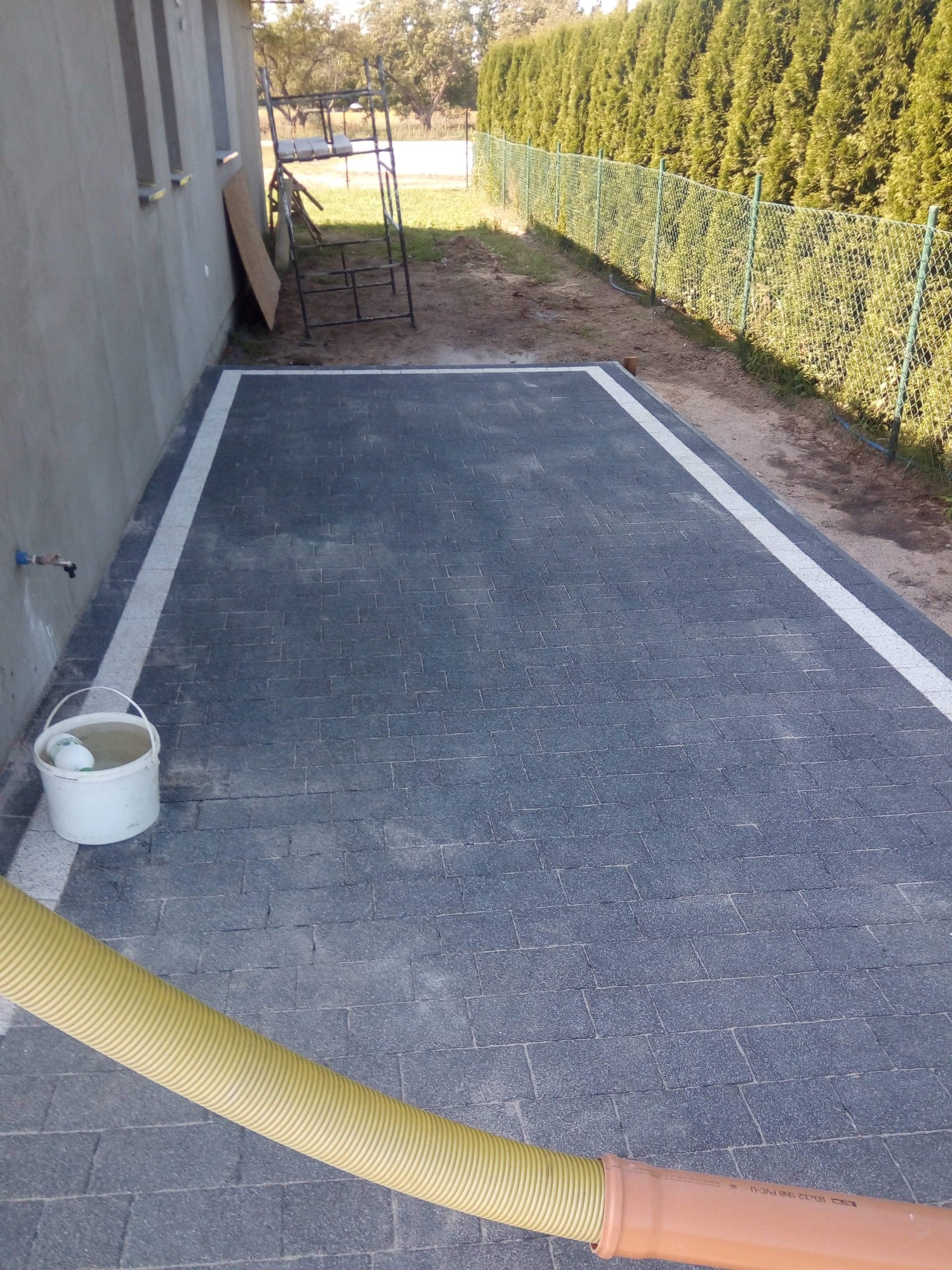 Ułożona kostka brukowa w kolorze grafitowym z białymi obrzeżami, widoczny żółty wąż i rura kanalizacyjna, ściana budynku i drabina w tle.