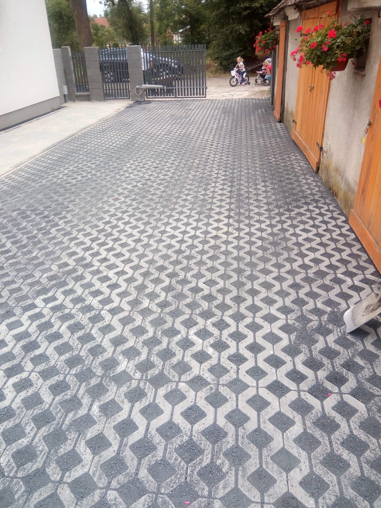 Szara kostka brukowa ułożona we wzór zygzakowaty, widoczne drewniane drzwi garażowe z wiszącymi czerwonymi kwiatami i brama w tle.