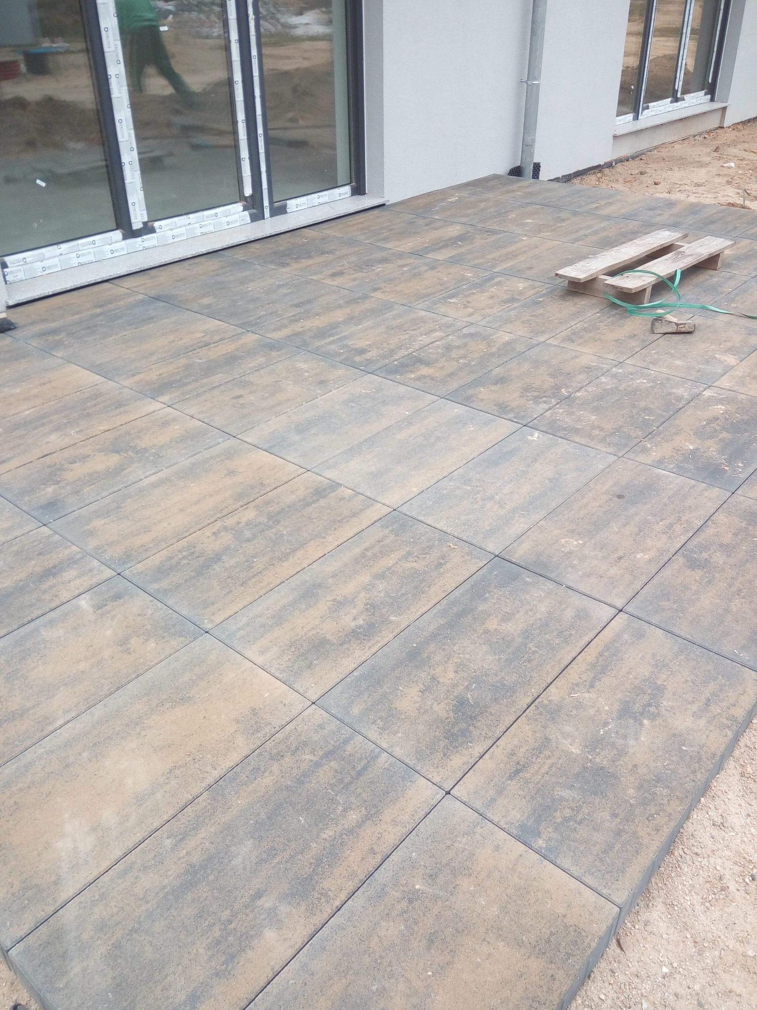 Wykończony taras z prostokątnej kostki brukowej w odcieniach szarości i beżu, z widocznym fragmentem budynku z dużymi oknami przesuwnymi i paletą oraz narzędziami w rogu.
