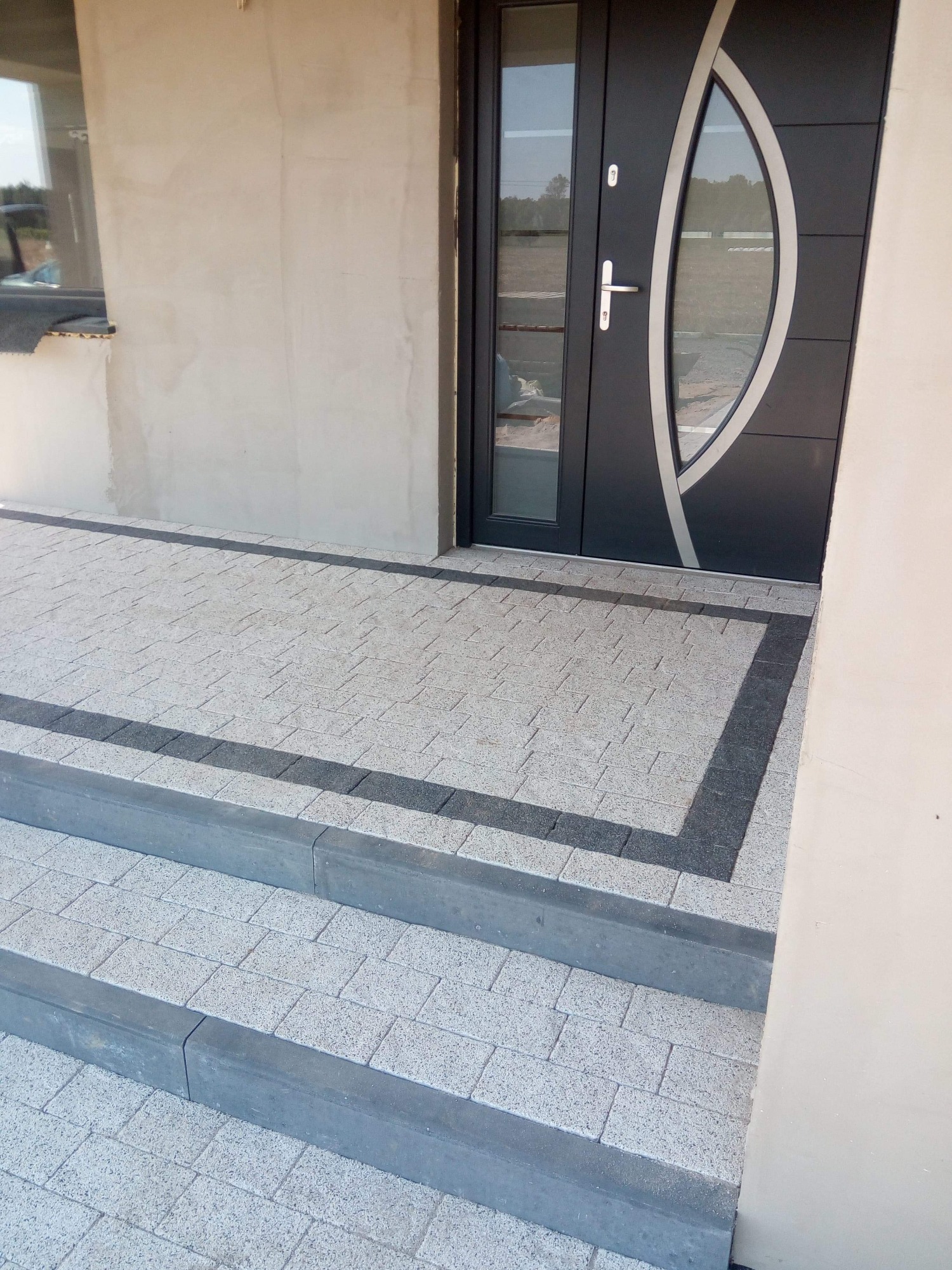 Ułożona kostka brukowa w odcieniach szarości z ciemniejszym obramowaniem prowadząca do nowoczesnych drzwi wejściowych z przeszkleniem.