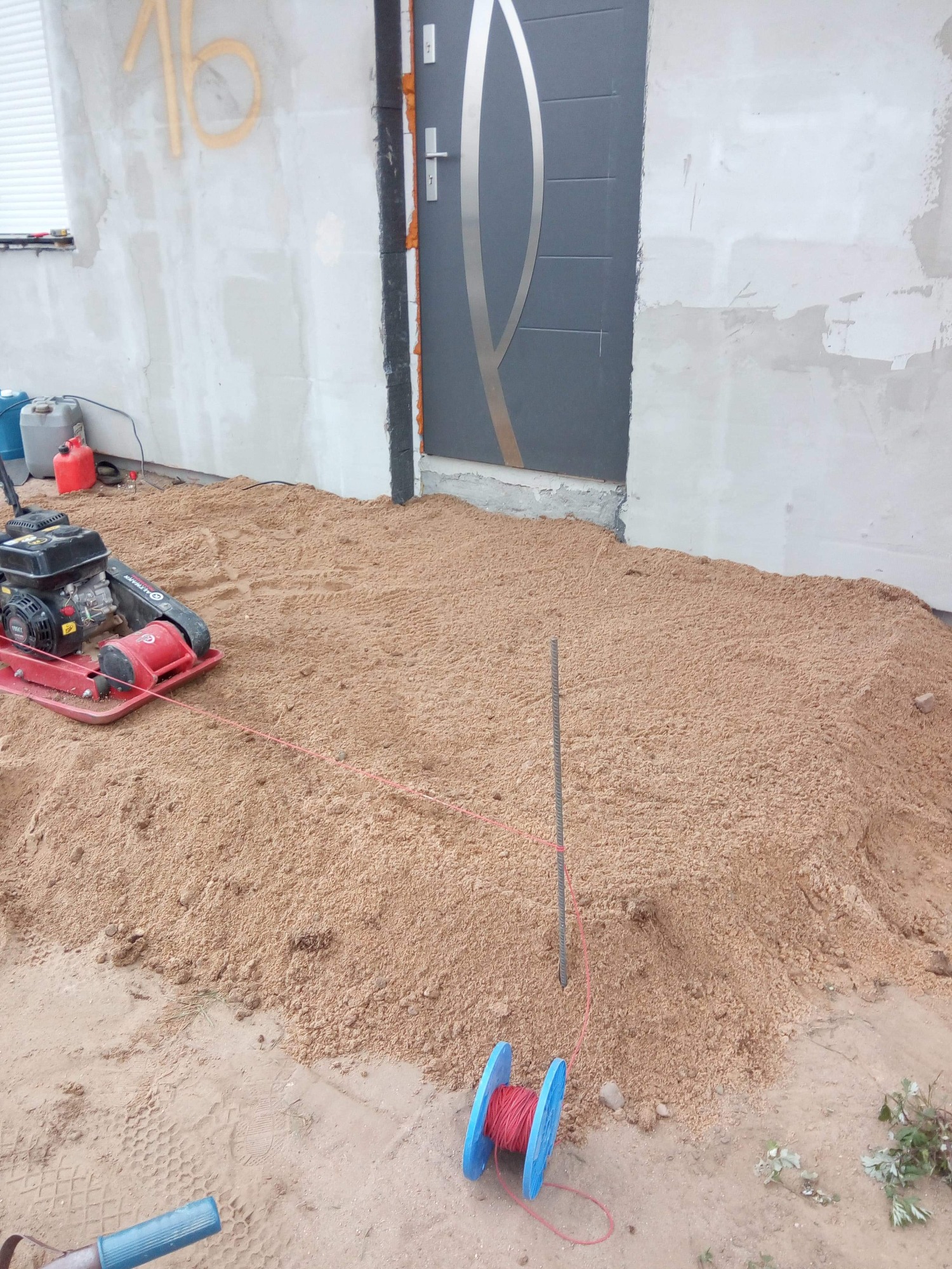 Przygotowanie podłoża pod kostkę brukową: ubijarka wibracyjna, wytyczony poziom i rozsypany piasek przed wejściem do domu w trakcie prac budowlanych.