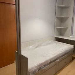 Białe łóżko z szufladami, materac zabezpieczony folią, zintegrowane z regałem z półkami i biurkiem, widoczne drewniane drzwi szafy przesuwnej.