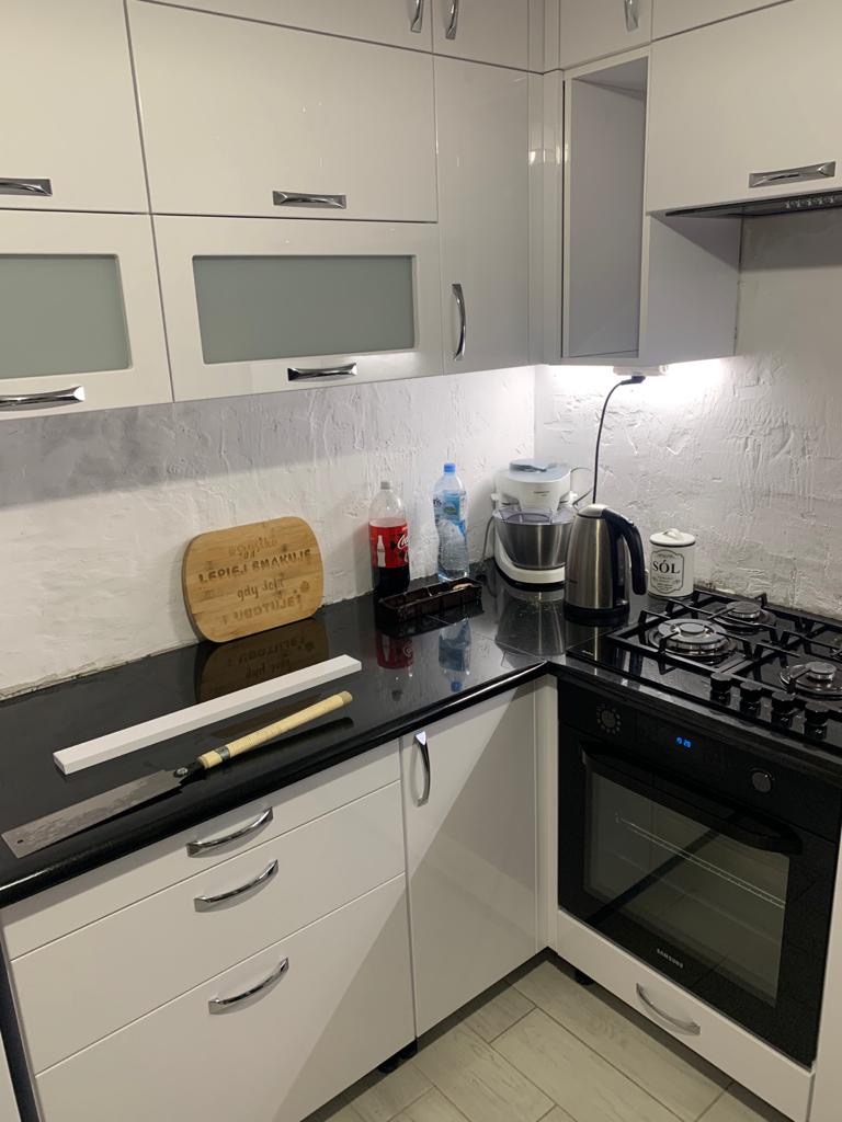 Biała, błyszcząca zabudowa kuchenna z czarnym blatem, na którym leżą narzędzia stolarskie: piła japońska i listwa przypodłogowa. Widoczna płyta gazowa i piekarnik. Na ścianie dekoracyjna deska...