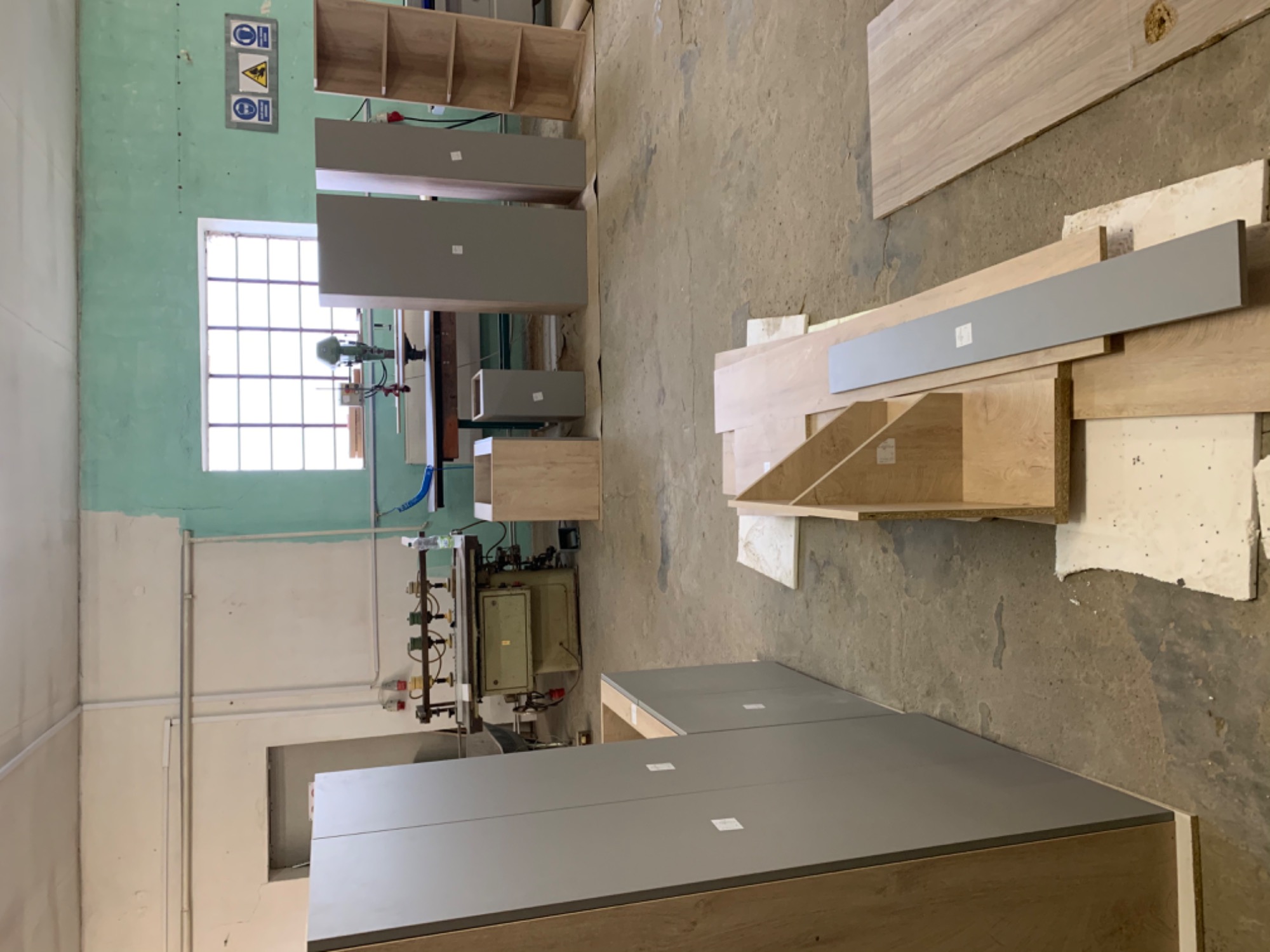 Elementy meblowe w warsztacie: szare fronty, drewniane półki i blaty, maszyna stolarska w tle, widok z góry.