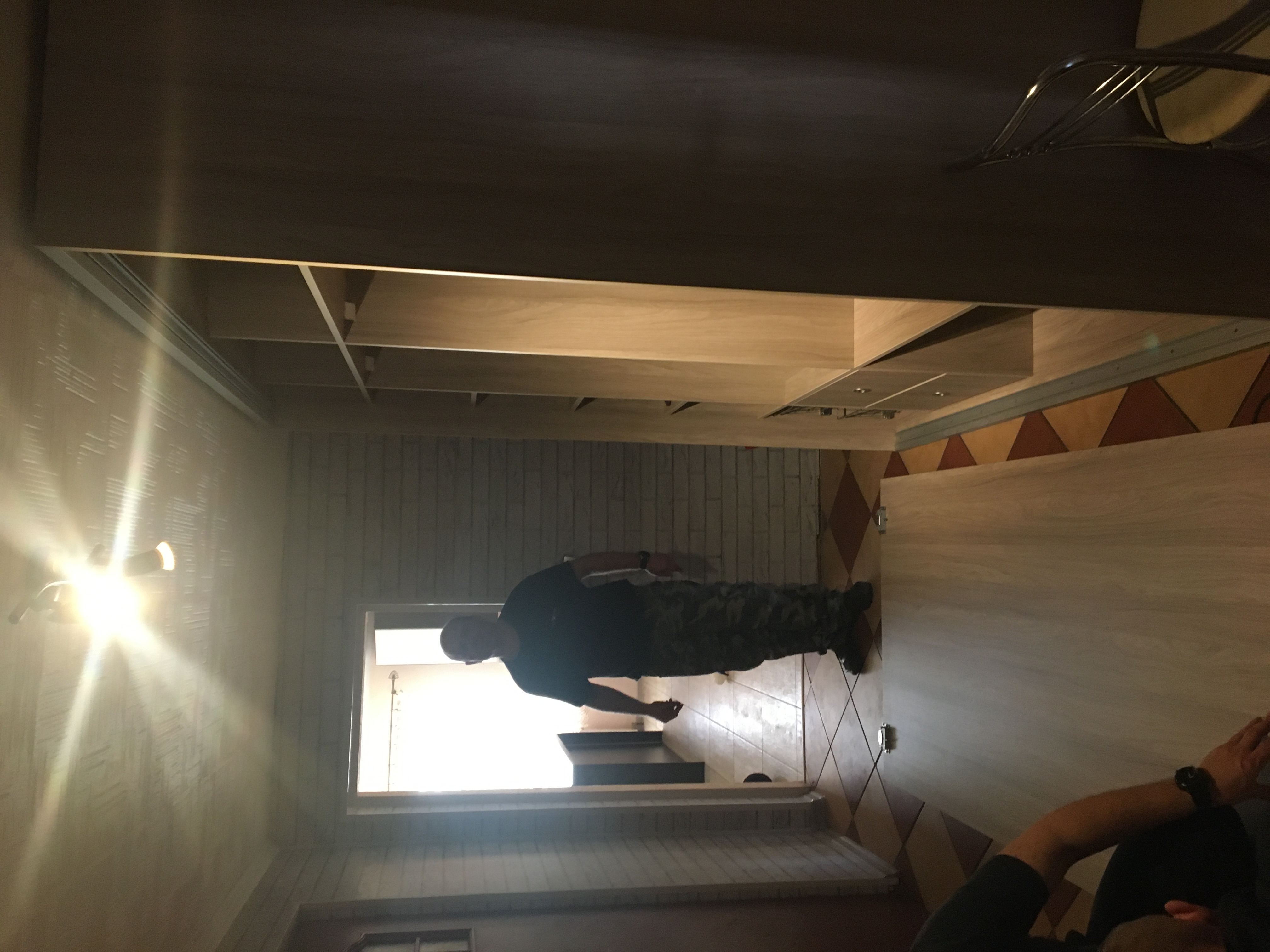 Instalacja jasnej, drewnianej szafy z półkami w pomieszczeniu z ceglaną ścianą i wzorzystą podłogą. Widoczny monter w czarnej koszulce i spodniach moro w trakcie pracy.