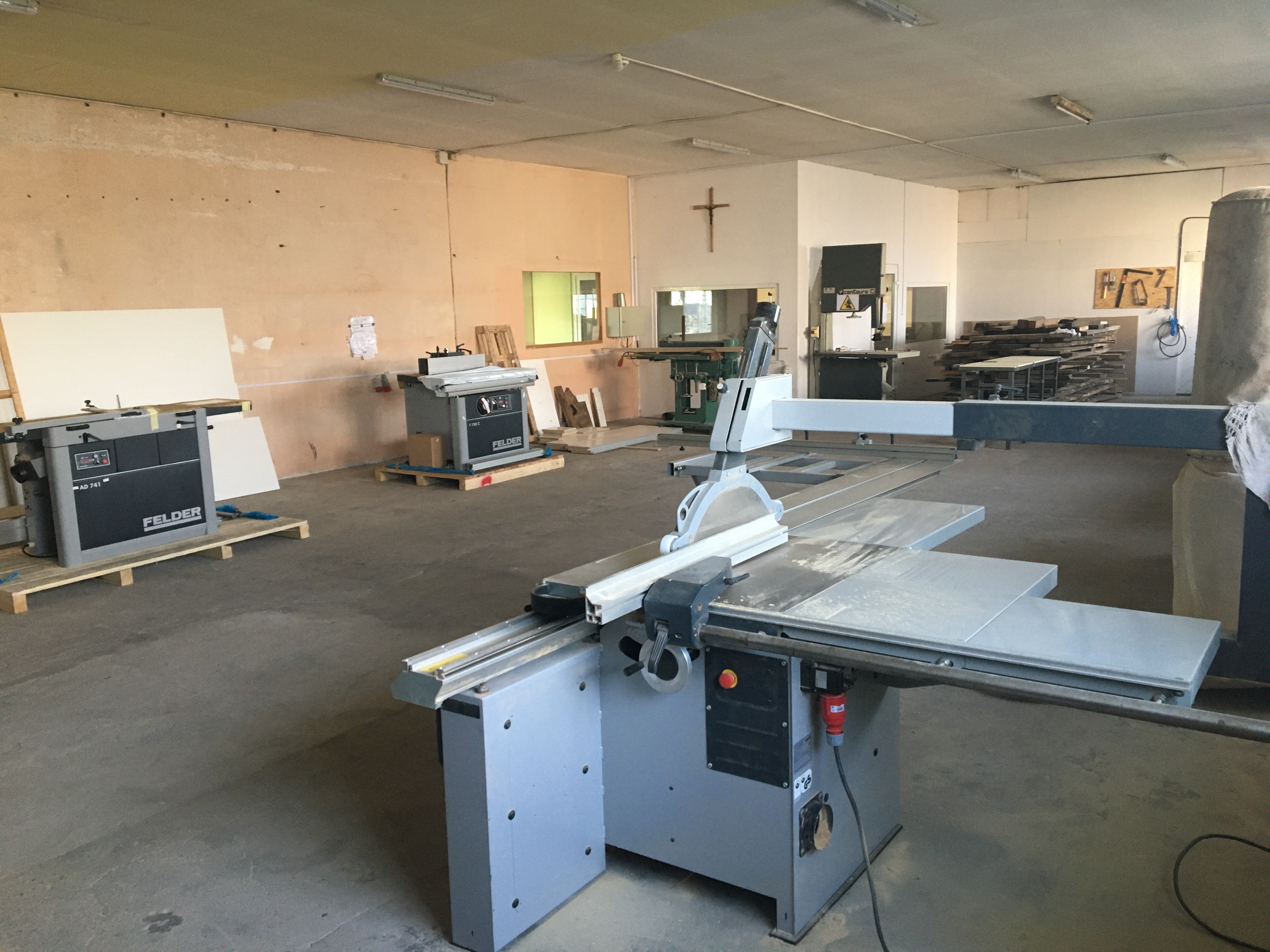 Warsztat stolarski z piłą formatową na pierwszym planie, inne maszyny do obróbki drewna w tle, krzyż na ścianie.