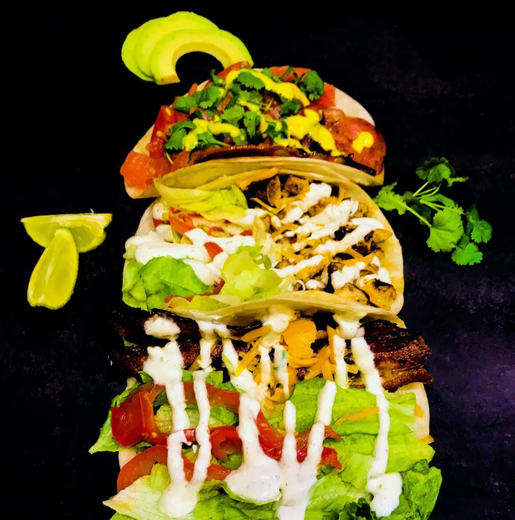 Trzy taco ułożone jeden na drugim na czarnym tle, z awokado i limonką jako dekoracją, widoczne różne rodzaje mięs, warzyw i sosów.