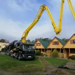 Czarny samochód ciężarowy z żółtym wysięgnikiem pompy do betonu na tle żółtych domków z zielonymi dachami, widoczne elementy zbrojenia i świeżo wylany beton.