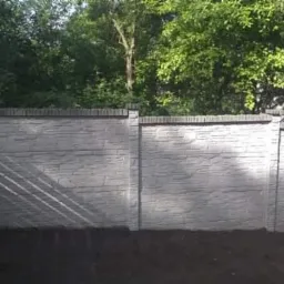 Fragment ogrodzenia betonowego z fakturą kamienia, zwieńczonego elementami imitującymi cegłę, na tle zieleni drzew.