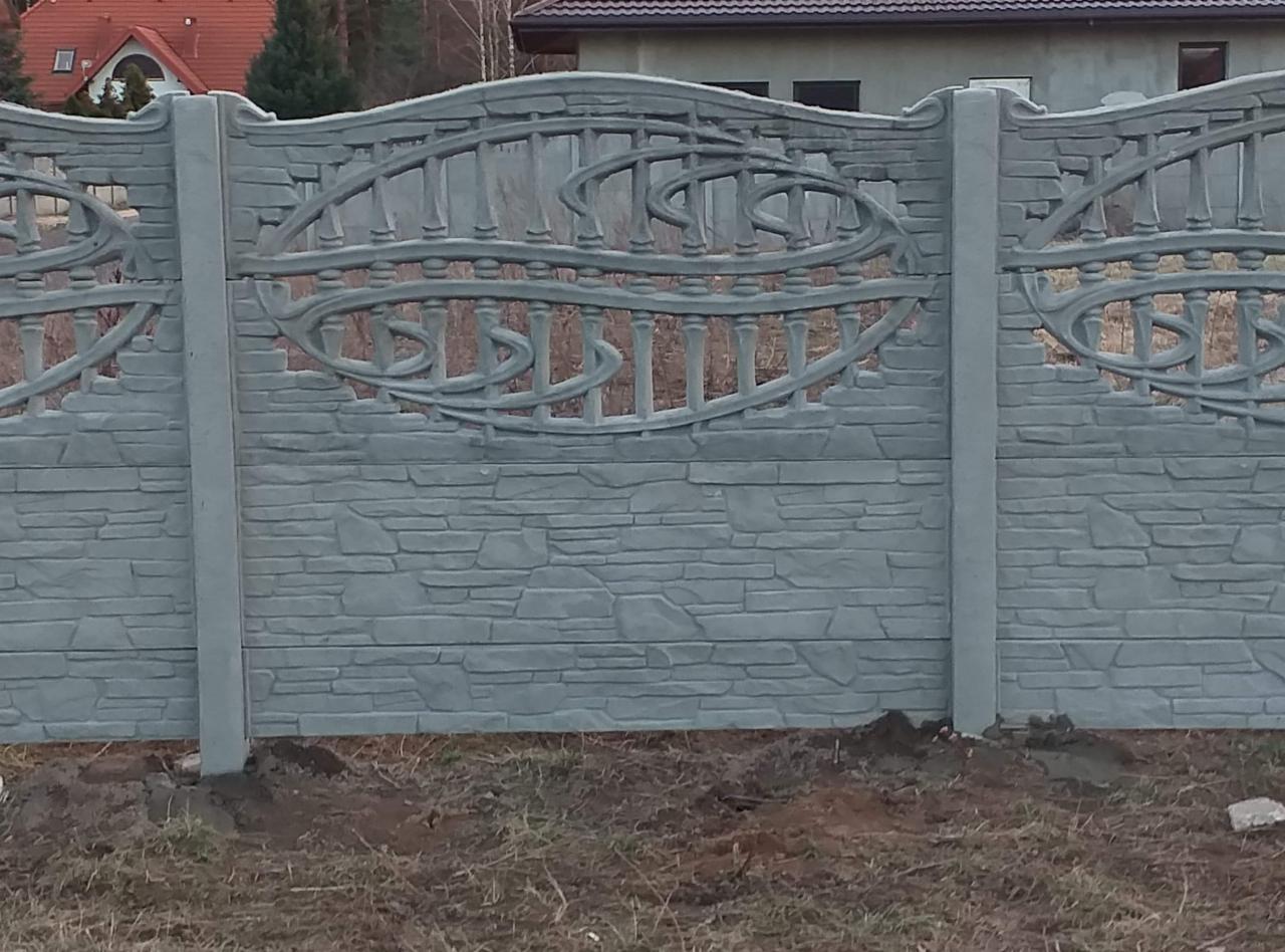 Szary, prefabrykowany fragment ogrodzenia z betonu imitującego kamień, z dekoracyjnym ażurem w górnej części, widziany w Opocznie po montażu, z widocznymi śladami ziemi po osadzeniu słupków.
