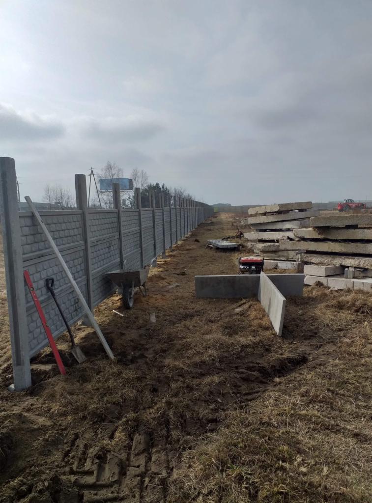 Długi, szary mur z prefabrykatów betonowych w trakcie montażu, widoczny poziomica, łopata, taczka oraz stosy płyt betonowych w tle, pochmurne niebo.