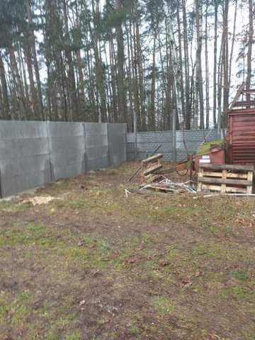 Wznoszenie ogrodzenia betonowego z prefabrykatów na działce z widocznymi drzewami w tle, częściowo ukończone, z paletami i narzędziami budowlanymi w pobliżu.