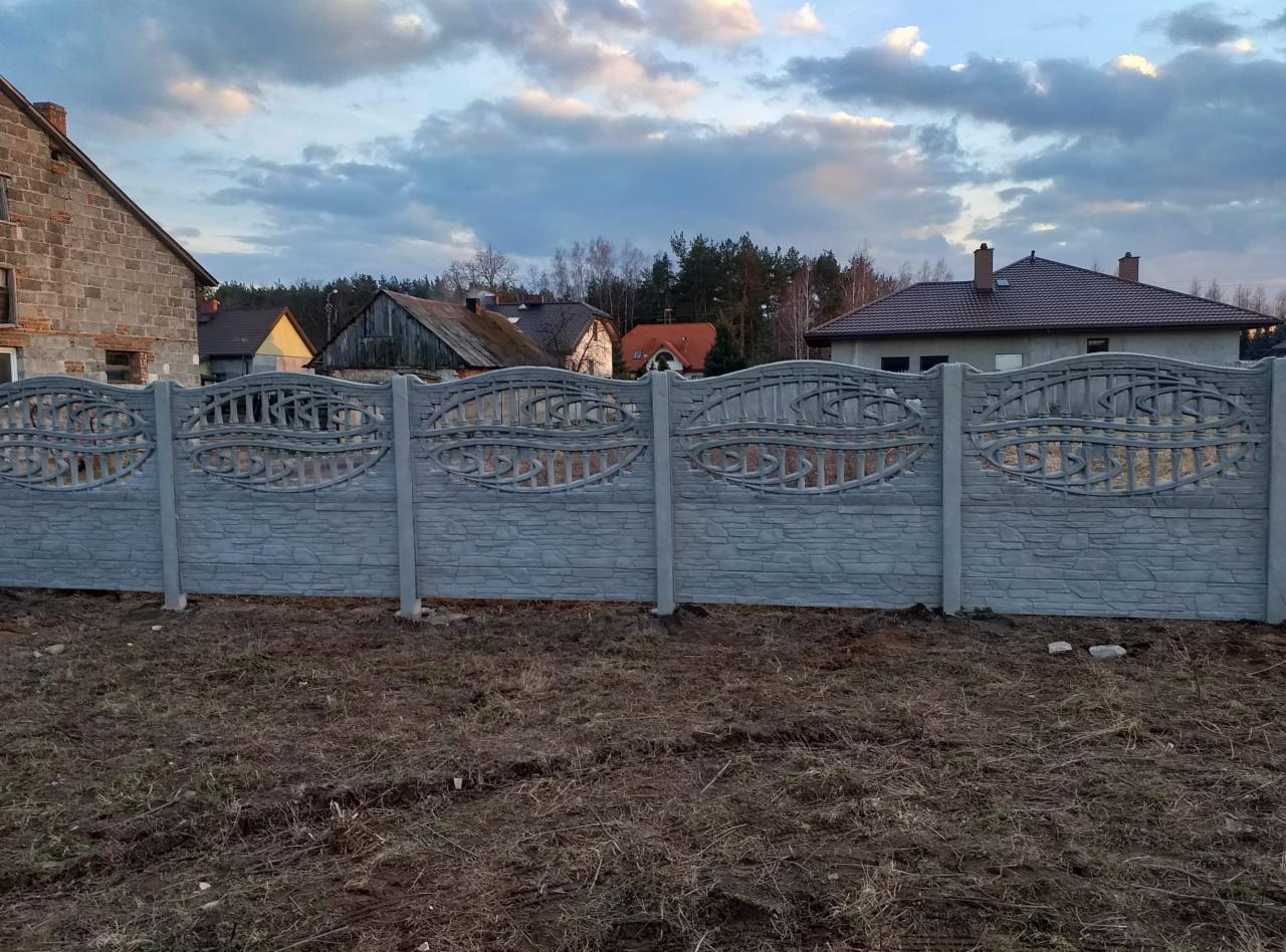 Szary, dekoracyjny płot z betonu imitującego kamień, z ozdobnymi ażurowymi wstawkami, na tle wiejskich domów i pochmurnego nieba.
