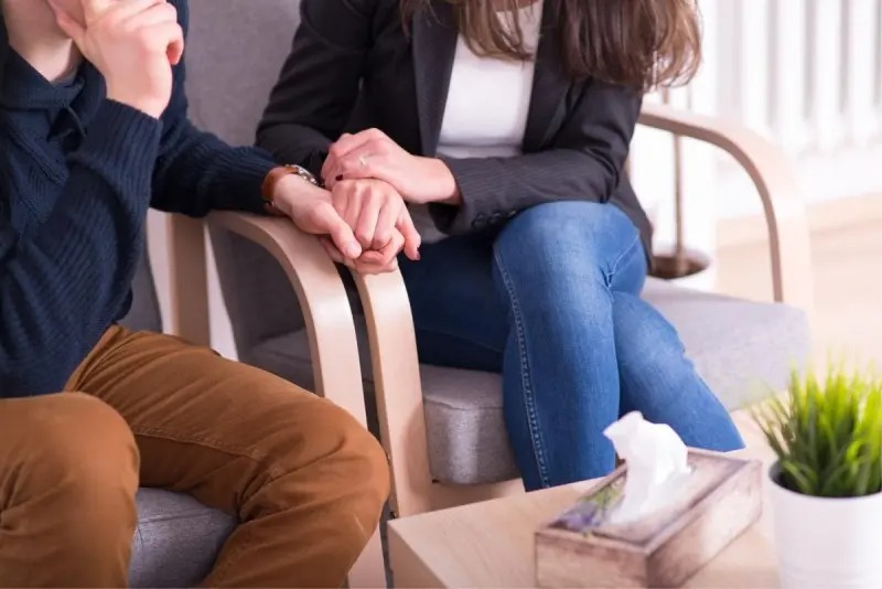 Para siedząca na fotelach trzymająca się za ręce, na stoliku pudełko chusteczek i roślina doniczkowa, kobieta w czarnej marynarce i jeansach, mężczyzna w brązowych spodniach i granatowym swetrze.