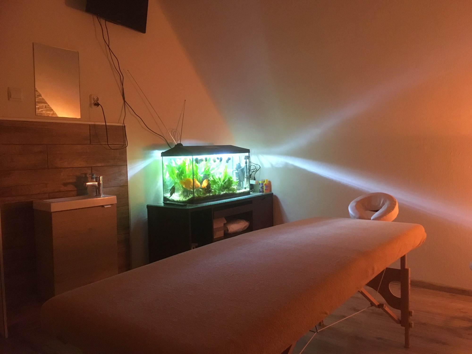 Pusta kozetka z zagłówkiem w gabinecie, oświetlona ciepłym światłem, z akwarium z rybkami w tle, obok umywalka z lustrem i telewizor na ścianie.