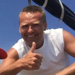 Uśmiechnięty mężczyzna w białej koszulce, dający kciuk w górę na tle żagla jachtu i błękitnego nieba.