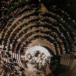 Widok z góry na amfiteatr podczas koncertu, scena z instrumentami i nagłośnieniem, publiczność siedząca na stopniach, oświetlenie punktowe.