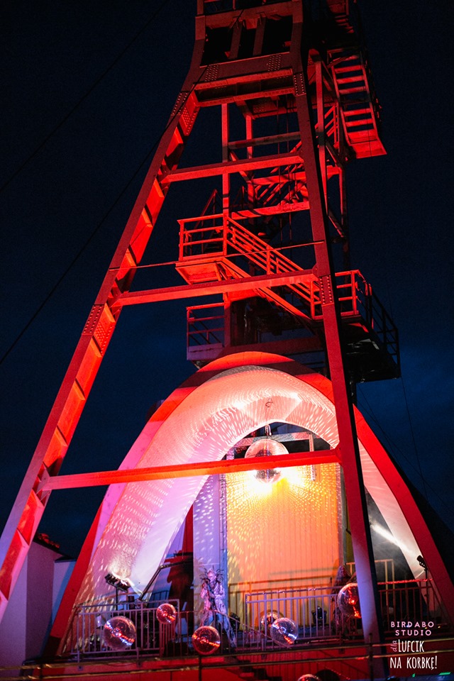 Metalowa konstrukcja wieży szybowej w czerwonym oświetleniu, udekorowana lustrzanymi kulami i białym materiałem, tworząc scenę eventową pod gołym niebem.