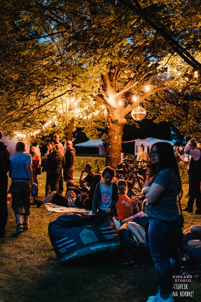 Plenerowa impreza podświetlona girlandami żarówek i dyskotekową kulą, uczestnicy siedzą na trawie i pufach, widoczny duży worek sako z logo firmy.