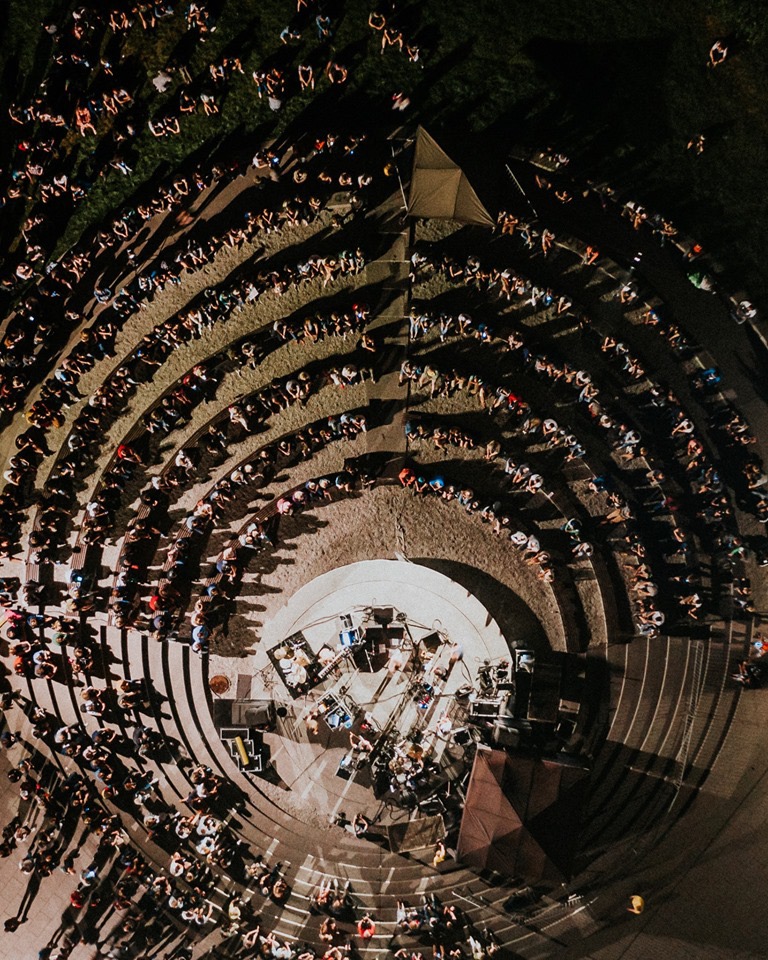 Widok z góry na amfiteatr podczas koncertu, scena z instrumentami i nagłośnieniem, publiczność siedząca na stopniach, oświetlenie punktowe.