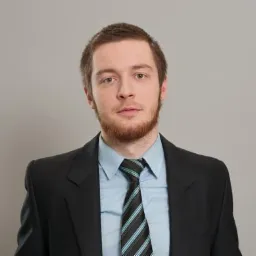 Portret młodego mężczyzny z rudą brodą w garniturze i krawacie w paski na szarym tle.