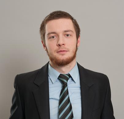 Portret młodego mężczyzny z rudą brodą w garniturze i krawacie w paski na szarym tle.