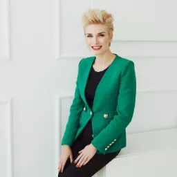 Uśmiechnięta kobieta w szmaragdowej marynarce z ozdobnymi guzikami, siedząca na białej komodzie na tle ściany z dekoracyjnymi panelami.