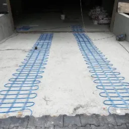 Montaż niebieskich kabli grzewczych na betonowej posadzce garażu przed wylaniem wylewki, widoczna zwinięta rolka kabla i skrzynki z materiałami w tle.