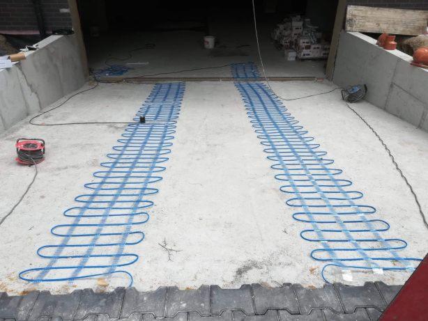 Montaż niebieskich kabli grzewczych na betonowej posadzce garażu przed wylaniem wylewki, widoczna zwinięta rolka kabla i skrzynki z materiałami w tle.