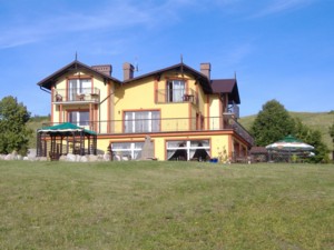 Żółty, dwupiętrowy budynek z balkonami i brązowym dachem, położony na wzgórzu, otoczony zielonym trawnikiem i drzewami, z widocznymi dwoma altanami ogrodowymi w tle błękitne niebo.