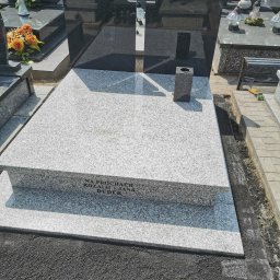 KAM-TAX - Nowoczesny nagrobek z jasnego granitu z ciemnym krzyżem w tle. Widoczne inskrypcje i otoczenie cmentarza. Minimalistyczny design.