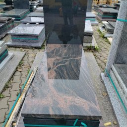 KAM-TAX - Polerowany, czarny nagrobek z brązowymi żyłkami, zabezpieczony taśmą. W tle inne nagrobki i elementy kamienne, na placu kamieniarskim.