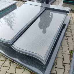 KAM-TAX - Szare, błyszczące płyty nagrobkowe z ciemną obwódką, ułożone na kostce brukowej. Na jednej z płyt odbija się sylwetka osoby i fragment drzewa.