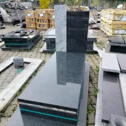 Gotowy nagrobek z ciemnego granitu z jasnoszarą płytą pionową, stojący na zewnątrz na placu z kostki brukowej, w tle drzewa i budynki.