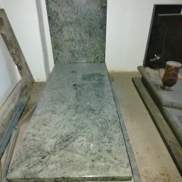 KAM-TAX DARIUSZ WIŚNIEWSKI - Jasnoszary nagrobek z granitu o wyraźnym wzorze, ustawiony w pomieszczeniu. Widoczne elementy innego nagrobka w tle.