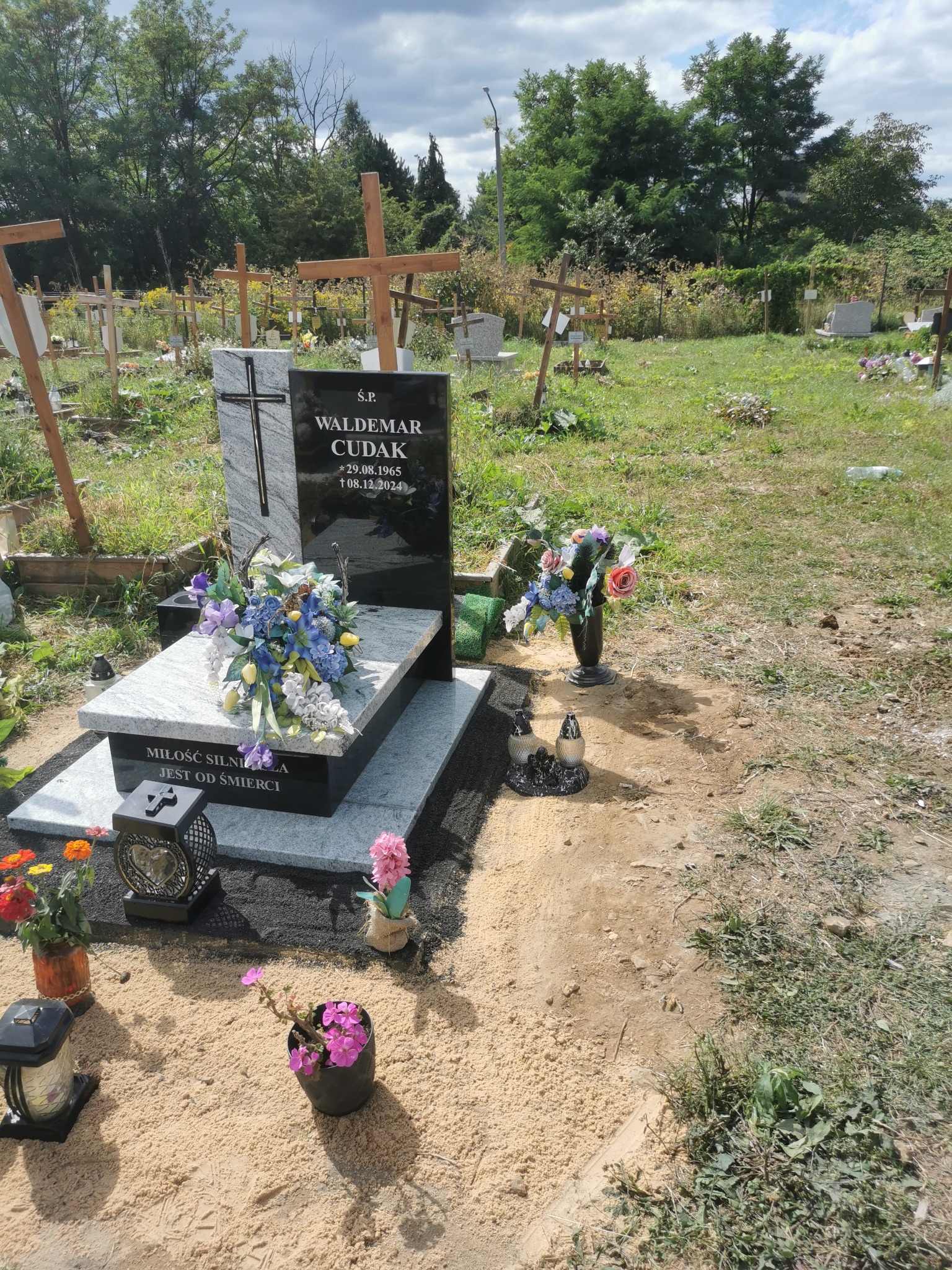 Granitowy nagrobek z czarną tablicą, ozdobiony kwiatami i lampionami na cmentarzu. W tle widoczne krzyże i zieleń.