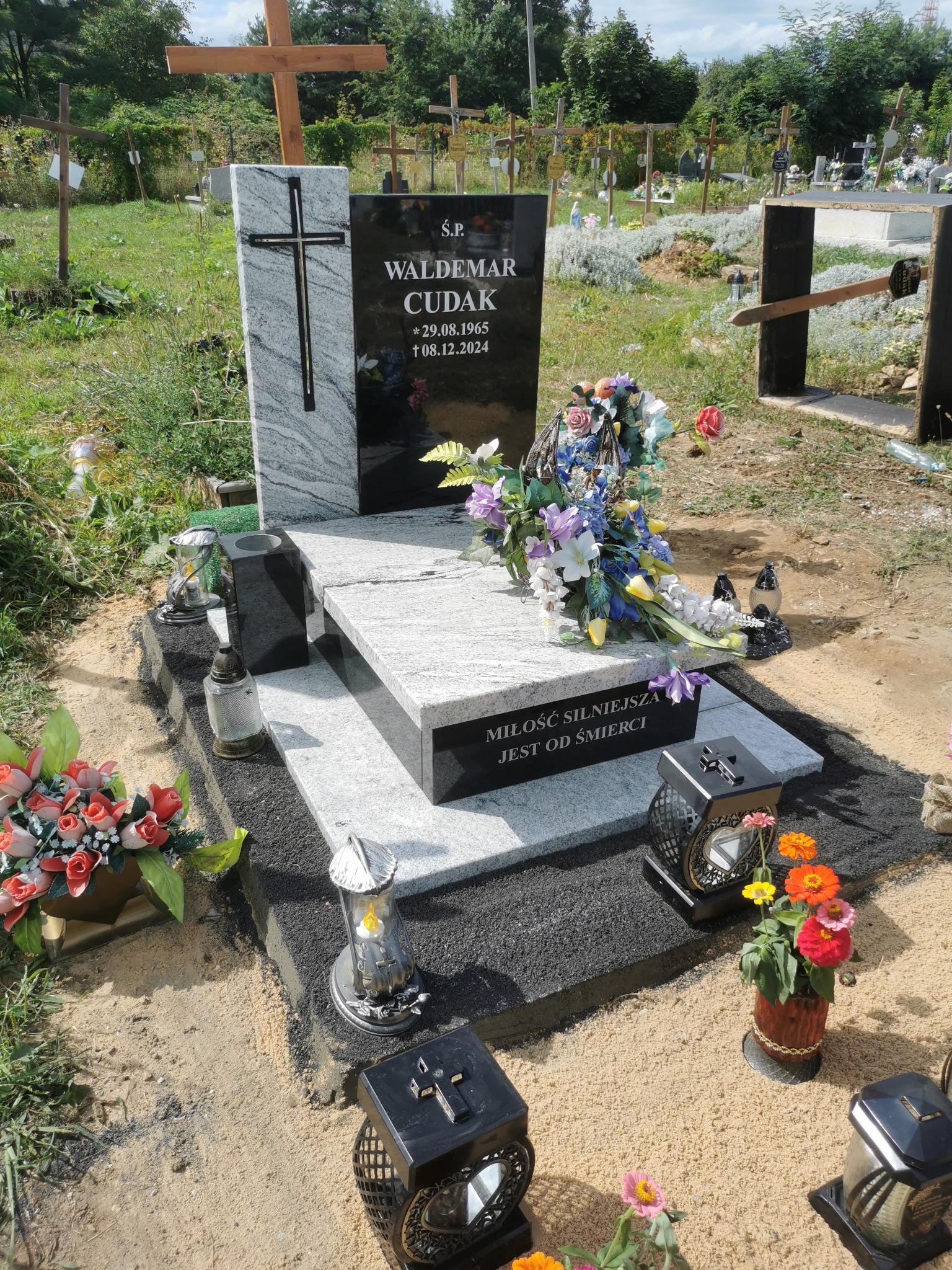 Granitowy nagrobek z krzyżem i inskrypcją na cmentarzu, otoczony kwiatami i zniczami, w tle inne groby i drzewa.