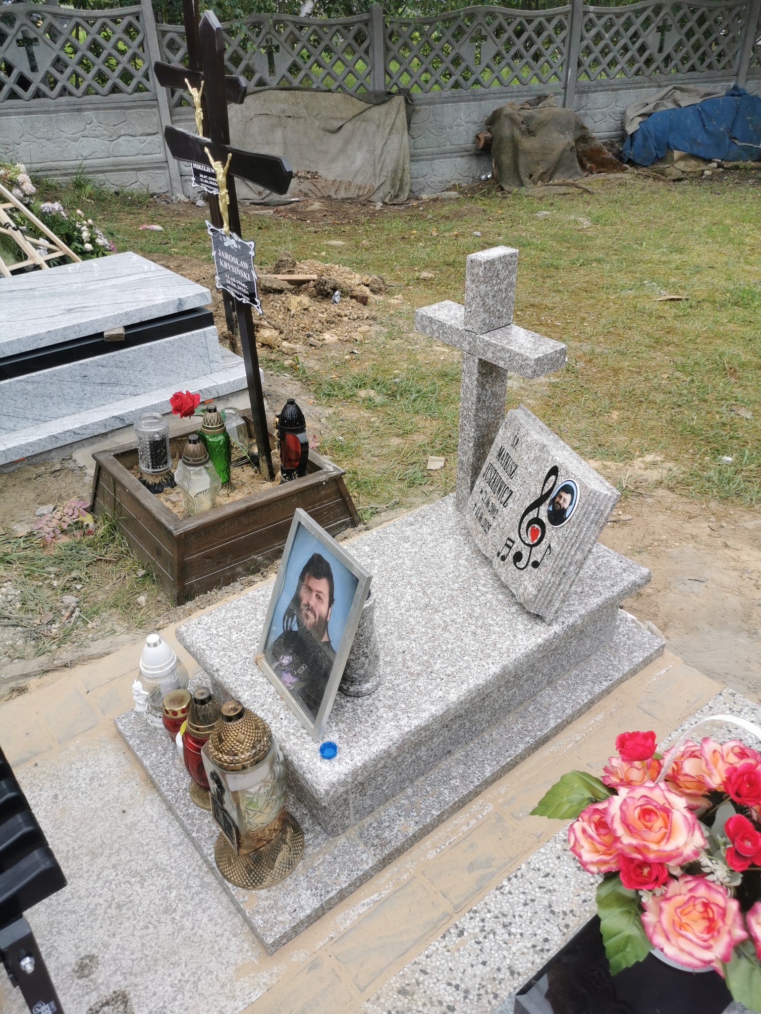 Jasnoszary nagrobek z krzyżem, zdjęciem zmarłego i motywami muzycznymi. Otoczenie: kwiaty, znicze, drewniana skrzynka. W tle betonowy płot i zieleń.