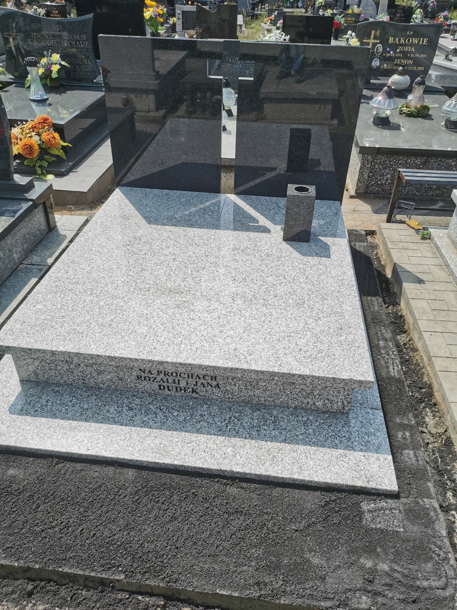 Nowoczesny nagrobek z jasnego granitu z ciemnym krzyżem w tle. Widoczne inskrypcje i otoczenie cmentarza. Minimalistyczny design.