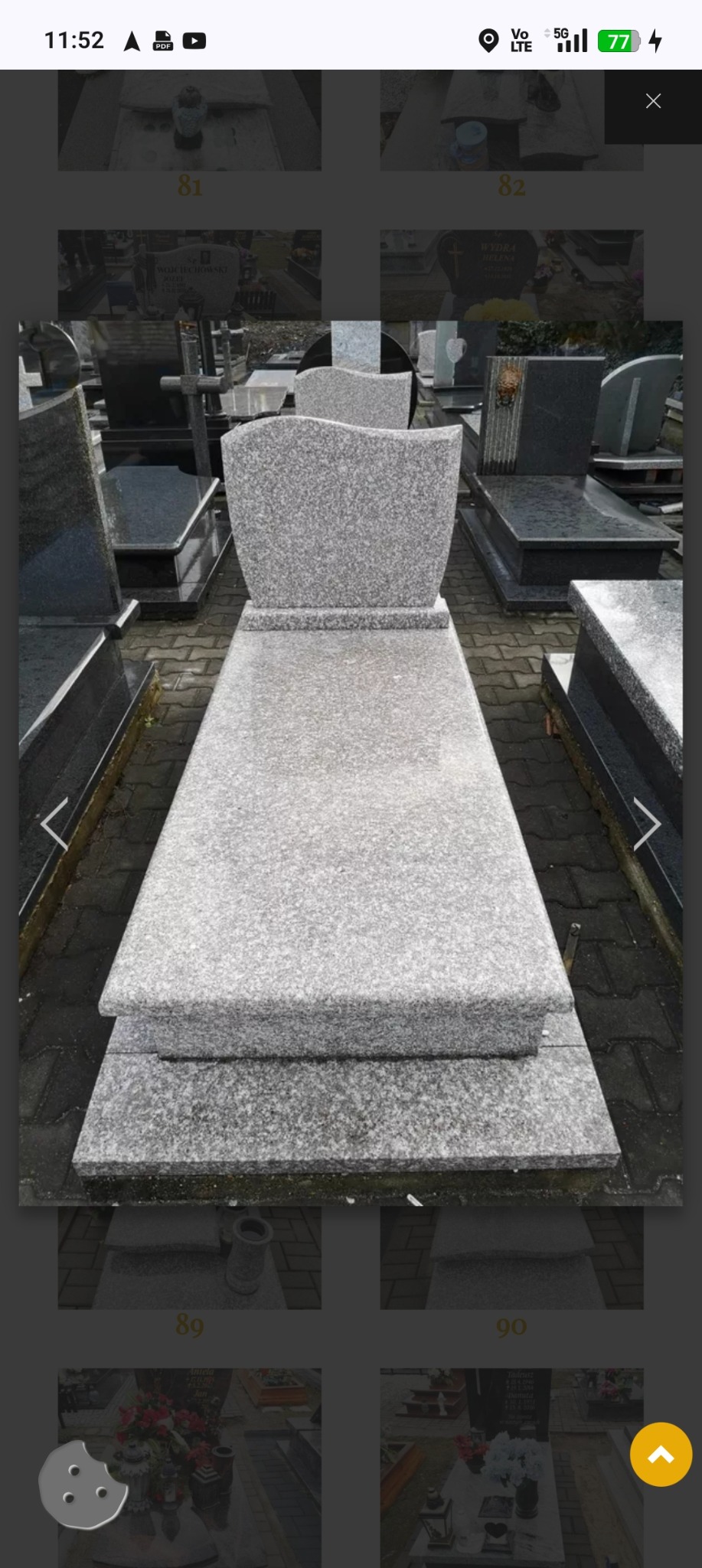 Jasnoszary, granitowy nagrobek pojedynczy z falistą górną krawędzią, na tle innych nagrobków. Widok z góry, na zewnątrz.