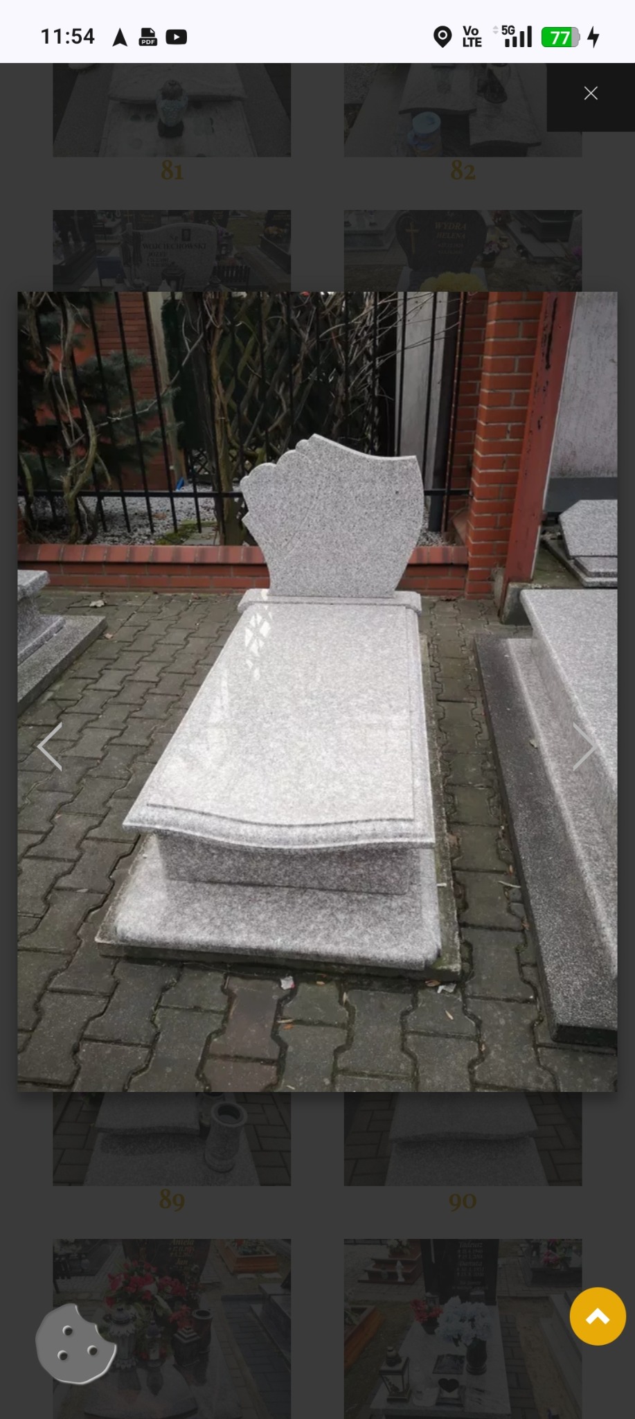 Jasnoszary, granitowy nagrobek z lekko zaokrąglonymi krawędziami, z widocznym obrysem płyty nagrobnej na kostce brukowej. W tle mur z czerwonej cegły.