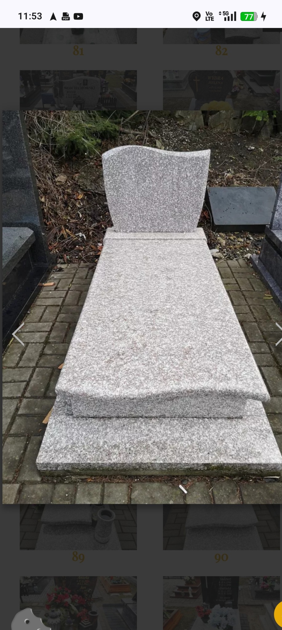 Jasnoszary, granitowy nagrobek z falistym zwieńczeniem, ustawiony na cmentarzu. Widoczne kostki brukowe i fragmenty innych nagrobków.