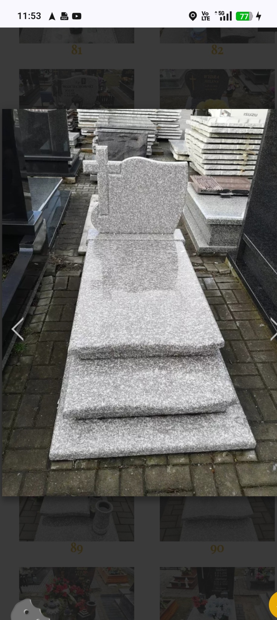 Jasnoszary, granitowy nagrobek z krzyżem, ustawiony na trzech stopniach, na tle innych nagrobków i kostki brukowej. Widok z przodu, lekko z góry.