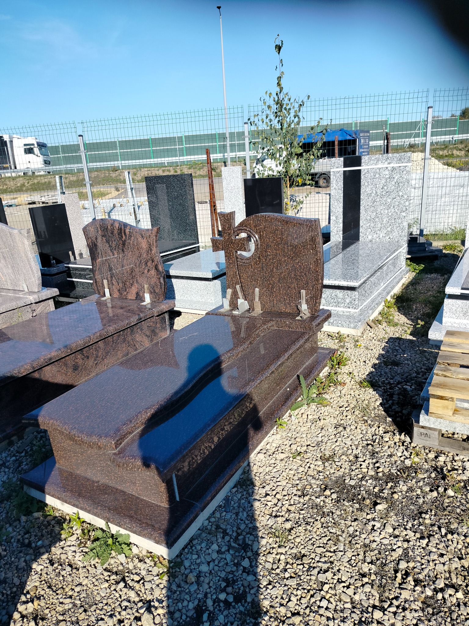 Brązowy, polerowany nagrobek z krzyżem, ustawiony na żwirowym podłożu, otoczony innymi nagrobkami i zielenią. Widoczne ogrodzenie i pojazd w tle.