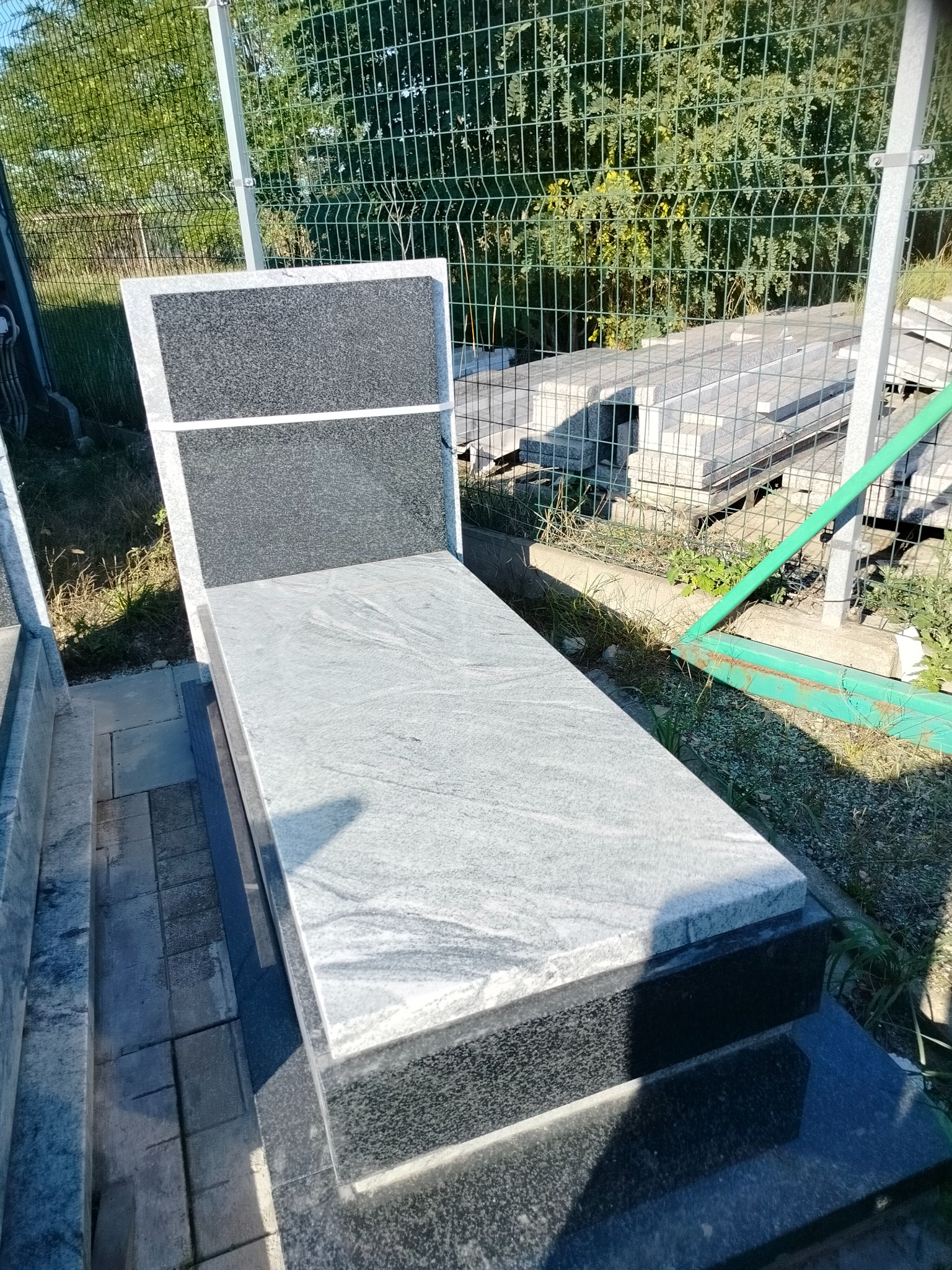 Jasnoszary nagrobek z czarnymi elementami na zewnątrz, widoczny na tle zieleni i ogrodzenia. Nagrobek wykonany z kamienia z wyraźną fakturą.