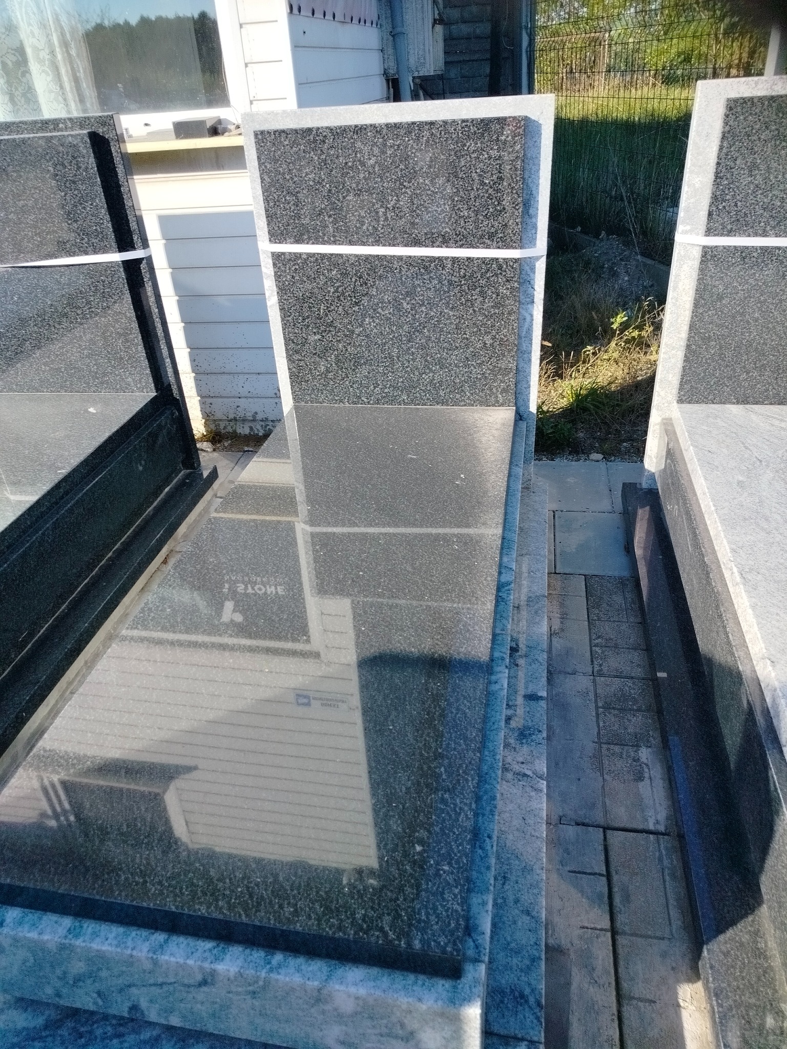 Ciemny, polerowany nagrobek z szarego kamienia z delikatnym wzorem, zabezpieczony białą taśmą, ustawiony obok innych nagrobków, na zewnątrz.